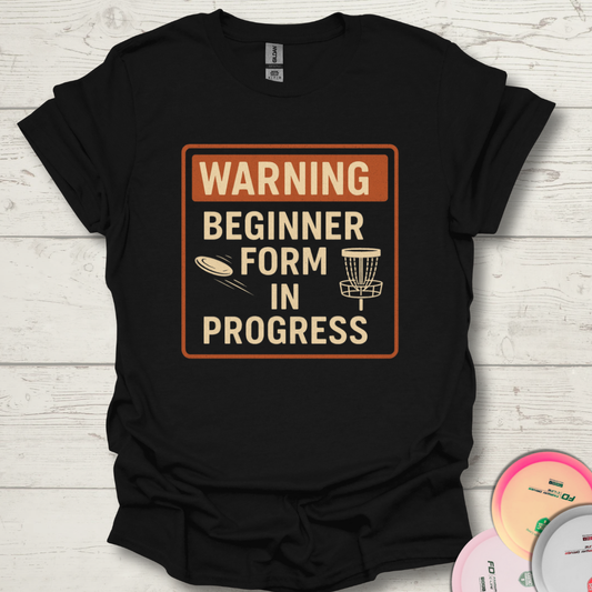 Warning Beginner Form In Progress funny disc golf t-shirt. Unisex funny beginner disc golf shirt on wooden background.